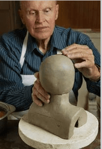 An elderly man sculpts a clay bust.